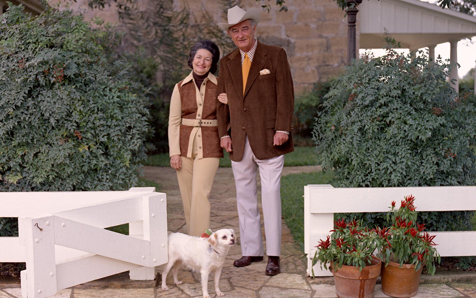 Lady Bird and Lyndon Johnson in Western wear with a small white dog, Yuki, standing by a low, white wooden fence in front of a ranch house.